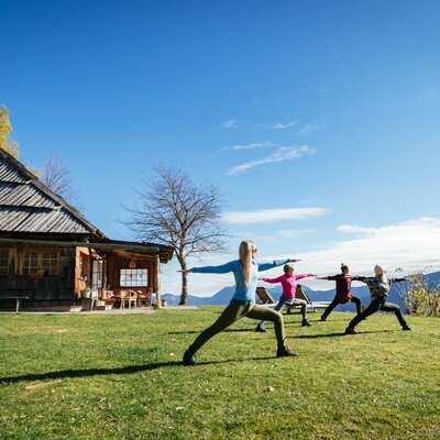 Eine Gruppe von Personen macht Yoga-Übungen auf einer Wiese vor einer traditionellen Holzhütte mit Bergkette im Hintergrund. | © Urlaub am Bauernhof Kärnten/ Daniel Gollner