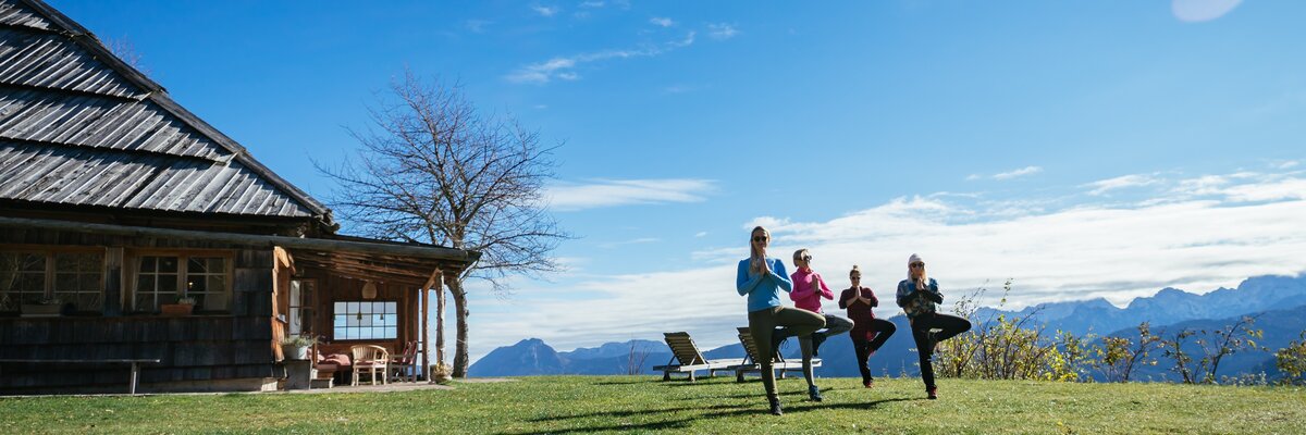 Eine Gruppe von Menschen praktiziert Yoga auf einer sonnigen Wiese vor einer Holzhütte mit Panoramablick auf die Berge. | © Urlaub am Bauernhof Kärnten/ Daniel Gollner