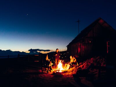 Abendfeuer vor alter Holzhütte, erhellt von Flammen in der Nacht, umgeben von Berglandschaft und Sternenhimmel. | © Urlaub am Bauernhof Kärnten/ Daniel Gollner 