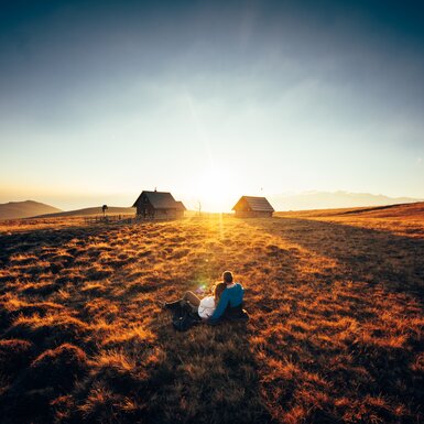 Ein romantischer Sonnenuntergang auf der Alm in Kärnten | © Urlaub am Bauernhof Kärnten/ Daniel Gollner Paar sitzt vor der Hütte im Gras und genießt den Sonnenuntergang auf der Alm | © Urlaub am Bauernhof Kärnten/ Daniel Gollner