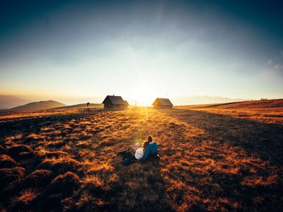 Ein romantischer Sonnenuntergang auf der Alm in Kärnten | © Urlaub am Bauernhof Kärnten/ Daniel Gollner Malerische Berghütten, von der Sonne durchflutete Wiese, zwei Personen genießen idyllischen Moment inmitten beeindruckender Landschaft. | © Urlaub am Bauernhof Kärnten/ Daniel Gollner
