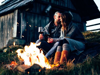 Ein Paar sitzt bei offenem Feuer in der Natur, umgeben von einer Holzhütte. Sie genießen den Abend mit einem Glas Wein, eingehüllt in eine Decke. | © Urlaub am Bauernhof Kärnten/ Daniel Gollner 