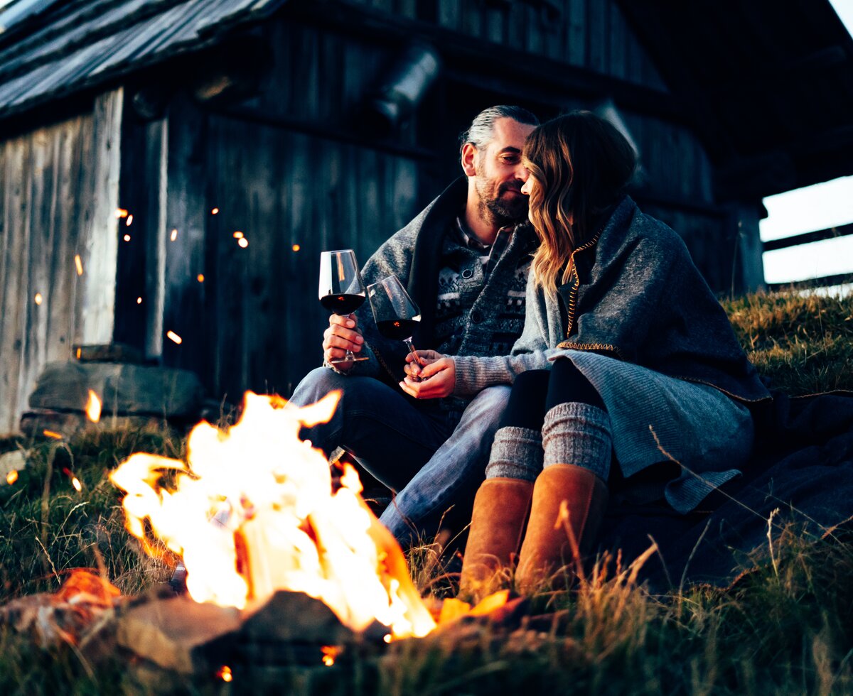 Ein Paar sitzt bei offenem Feuer in der Natur, umgeben von einer Holzhütte. Sie genießen den Abend mit einem Glas Wein, eingehüllt in eine Decke. | © Urlaub am Bauernhof Kärnten/ Daniel Gollner 