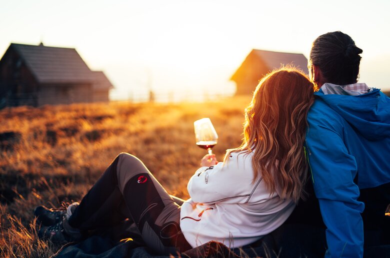 Ein junges Paar sitzt auf einer Wiese und genießt den Sonnenuntergang. Der Partner hält ein Glas Wein in der Hand, während sie gemeinsam den warmen Ausblick betrachten. | © Urlaub am Bauernhof Kärnten/ Daniel Gollner