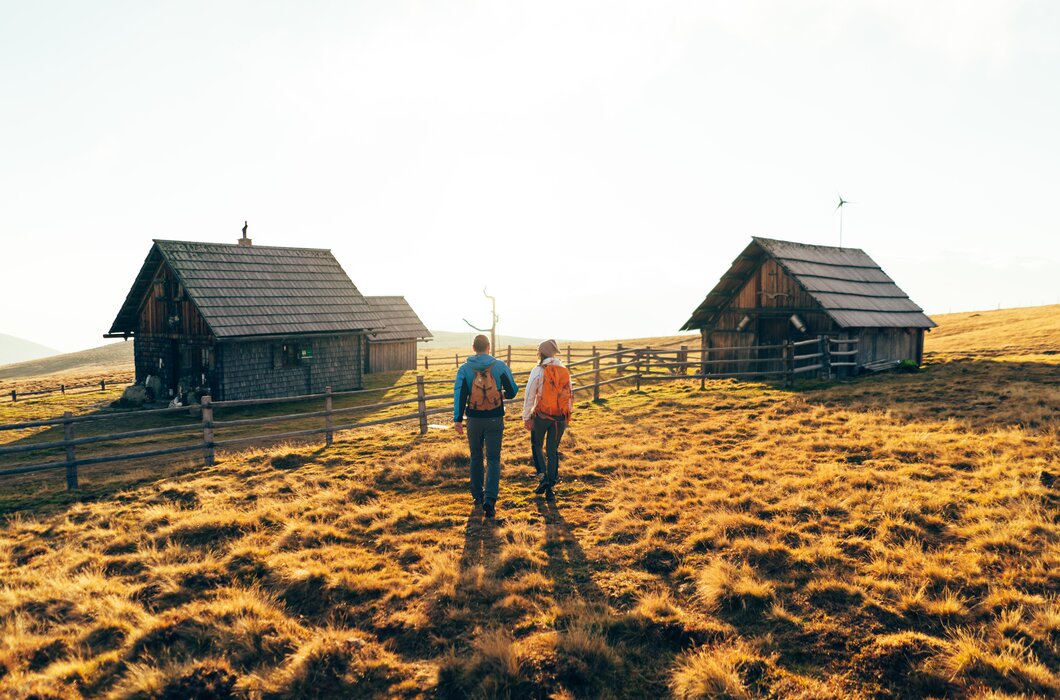 Zwei Wanderer stehen vor alten Holzgebäuden, von Zäunen umgeben, auf einer sonnenlichtdurchfluteten Wiese. Die Gebäude haben schiefergedeckte Dächer und sind in einem ländlichen Ambiente eingebettet.