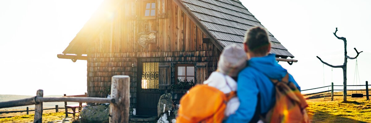 Traditionelles Holzhaus mit Reetdach, von Wanderern mit Rucksäcken besucht, inmitten einer Landschaft mit Zaun und Baum. Warmes, sonniges Licht umspielt die Szenerie.