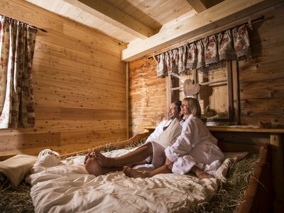 Ein gemütliches Holzhütten-Interieur mit einem Bett aus Naturmaterialien, auf dem eine verliebtes Paar entspannt beisammensitzt, umgeben von Dekorationen im rustikalen Stil. | © Martin Steinthaler / Urlaub am Bauernhof Kärnten