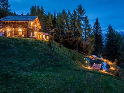 Premiumhütte Aussenaufnahme | © Günter Standl Ein beleuchtetes Holzblockhaus thront auf einem grünen Hügel, umgeben von dichten Nadelbäumen. Ein beleuchteter Steg führt zu einem kleinen See, der das Haus in seinem Wasser spiegelt. Die Atmosphäre ist gemütlich und idyllisch. | © Günter Standl
