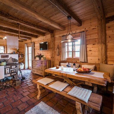 Gemütliches Holzinterieur mit rustikalem Esszimmertisch, einem Kaminofen und einer offenen Küche. Dekorative Details, wie Blumen und Naturalien, verleihen dem Raum eine charmante Atmosphäre. | © Günter Standl 