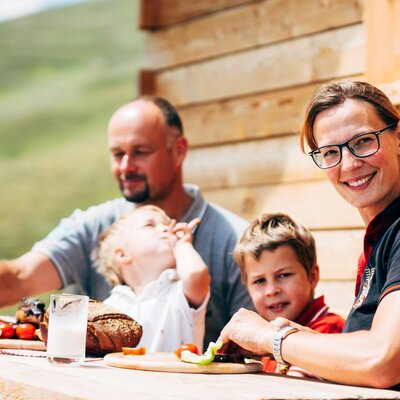 Eine vierköpfige Familie genießt gemeinsam ein Mahlzeit an einem Holztisch außerhalb eines Hauses. Sie lachen und scheinen in einer entspannten Umgebung zu sein. | © Urlaub am Bauernhof Kärnten / Daniel Gollner