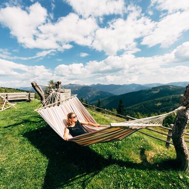 Person in bunter Hängematte genießt den Ausblick auf die grüne Berglandschaft und den bewölkten Himmel an einem sonnigen Tag. | © Urlaub am Bauernhof Kärnten / Daniel Gollner
