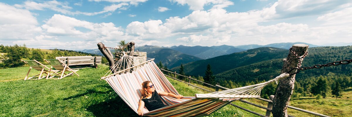 Person in bunter Hängematte genießt den Ausblick auf die grüne Berglandschaft und den bewölkten Himmel an einem sonnigen Tag. | © Urlaub am Bauernhof Kärnten / Daniel Gollner