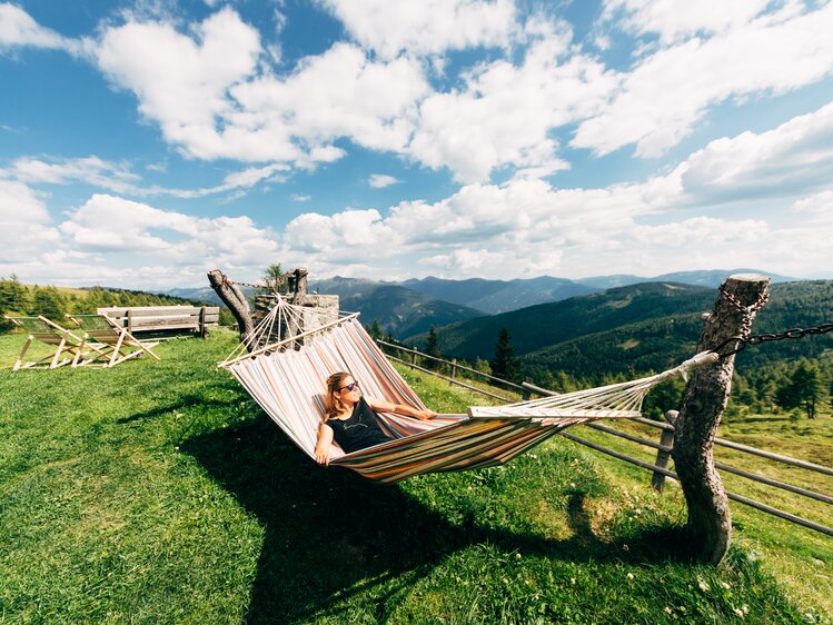 Person in bunter Hängematte genießt den Ausblick auf die grüne Berglandschaft und den bewölkten Himmel an einem sonnigen Tag. | © Urlaub am Bauernhof Kärnten / Daniel Gollner
