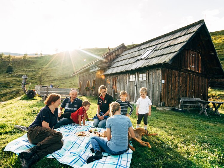 Eine Gruppe sitzt auf einer Decke in einem ländlichen Umfeld. Im Hintergrund ein altes Holzhaus mit Strohdach, umgeben von Wiesen und Hügeln. Die Menschen scheinen eine gesellige Picknick-Auszeit in der Natur zu genießen. | © Urlaub am Bauernhof Kärnten / Daniel Gollner