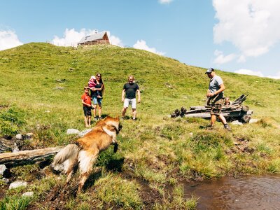 Wanderer mit Hund erkunden eine Berglandschaft mit Holzhütte auf einem grünen Hügel. Im Vordergrund ein Bachlauf, dahinter eine Gruppe Menschen, die mit einem Motorrad herumstehen. | © Urlaub am Bauernhof Kärnten / Daniel Gollner