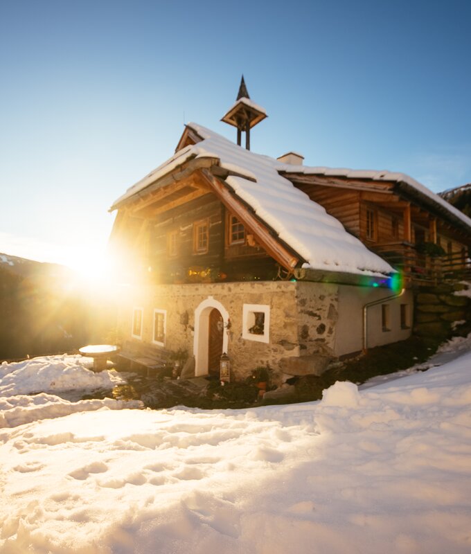 Altes, traditionelles Holzhaus mit Satteldach in verschneiter, bergiger Landschaft. Sonniges Licht erfüllt die Szene, verleiht dem Haus eine verträumte, idyllische Stimmung. | © Urlaub am Bauernhof Kärnten / Daniel Gollner