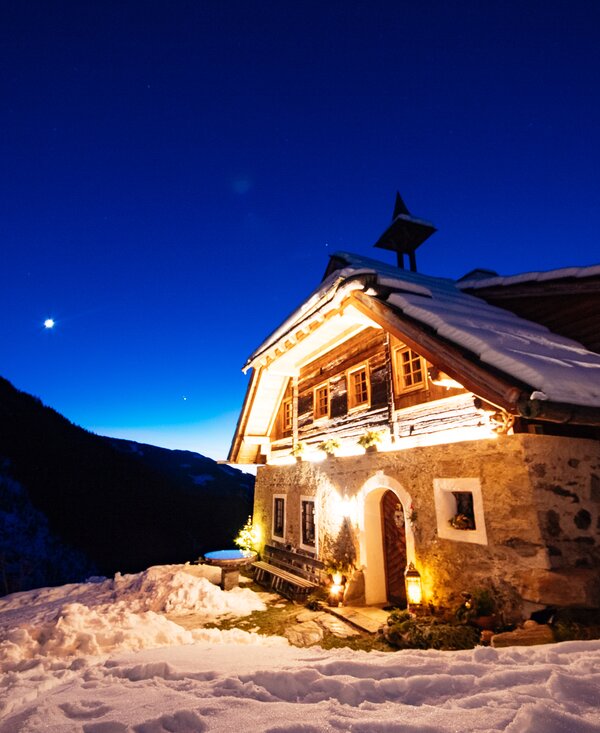Sandrisser Hütte bei Nacht | © Daniel Gollner / Urlaub am Bauernhof Kärnten  Holzhütte in verschneiter Berglandschaft bei Nacht, beleuchtet durch Fackelschein und Sterne am tiefblauen Nachthimmel. | © Daniel Gollner / Urlaub am Bauernhof Kärnten