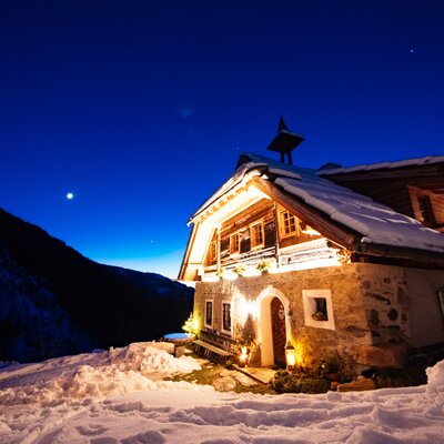 Sandrisser Hütte bei Nacht | © Daniel Gollner / Urlaub am Bauernhof Kärnten  Holzhütte in verschneiter Berglandschaft bei Nacht, beleuchtet durch Fackelschein und Sterne am tiefblauen Nachthimmel. | © Daniel Gollner / Urlaub am Bauernhof Kärnten