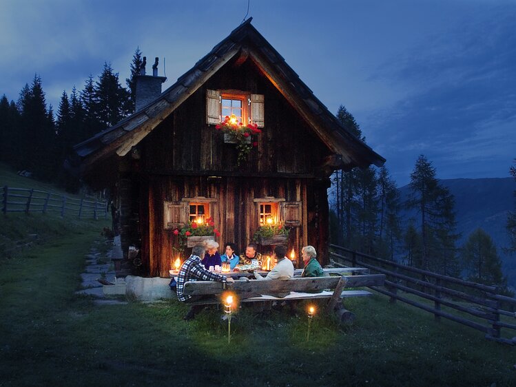 Eine urige Holzhütte bei Nacht, geschmückt mit Blumen und Lichterketten. Mehrere Personen sitzen gemütlich an einem Tisch, umgeben von der malerischen Waldlandschaft. | © Urlaub am Bauernhof Kärnten/ Ernst P. Prokop