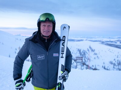 Skifigur in Winterkleidung, haltend Ski und Stöcke, vor verschneiter Berglandschaft mit Skiliften im Hintergrund. | © BRM / Mathias Prägant