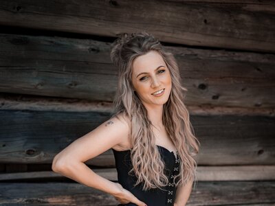 Junge Frau mit langen, blonden Haaren und Fringerock in Schwarztönen und leuchtendrosa. Sie steht lächelnd vor einer Holzwand. | © Lambauer Entertainment / Christoph Hatheuer
