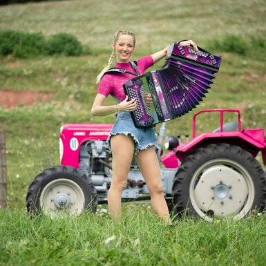 Eine jung aussehende Frau in pinker Kleidung spielt auf einem roten Traktor Akkordeon, umgeben von einer Wiese mit Blumen. | © Lambauer Entertainment / Christoph Hatheuer