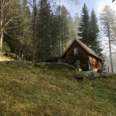 Ein baufälliges Holzhaus steht inmitten eines bewaldeten Berghanggeländes mit Moos und abgestorbenen Ästen. Im Vordergrund sind zwei Personen in Regenjacken zu erkennen. | © Urlaub am Bauernhof / Schell