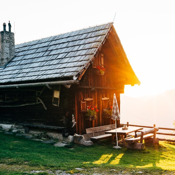 Altes, hölzernes Berghaus mit Strohdach und Schornstein, eingebettet in eine grüne Umgebung mit Holzbänken und Blumen. Die goldene Abendsonne taucht das Bild in ein warmes Licht. | © Urlaub am Bauernhof Kärnten / Daniel Gollner