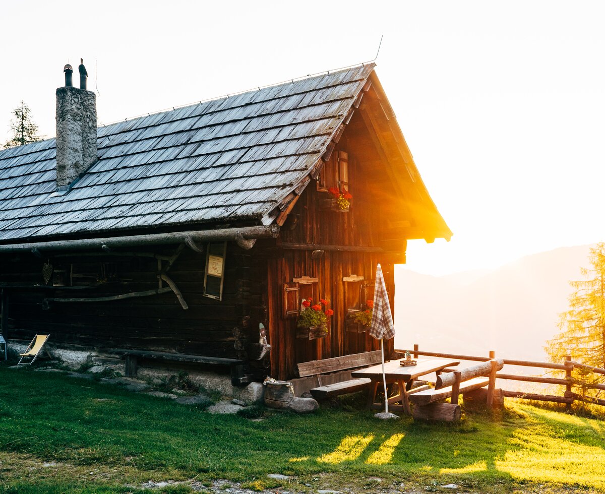 Altes, hölzernes Berghaus mit Strohdach und Schornstein, eingebettet in eine grüne Umgebung mit Holzbänken und Blumen. Die goldene Abendsonne taucht das Bild in ein warmes Licht. | © Urlaub am Bauernhof Kärnten / Daniel Gollner