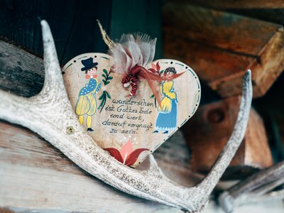 Ein bemaltes Herz an einem verwitterten Holzbrett, geschmückt mit Federn und einer bunten Blume. Darauf befindet sich eine handschriftliche Aufschrift in deutscher Sprache. | © Urlaub am Bauernhof Kärnten / Daniel Gollner