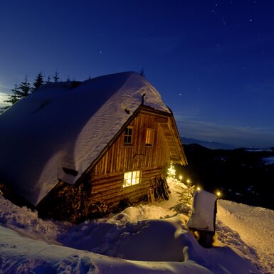 Eine verschneite Berghütte mit beleuchteten Fenstern und einer schneebedeckten Umgebung unter einem sternenübersäten Nachthimmel. | © Urlaub am Bauernhof/ Tom Lamm