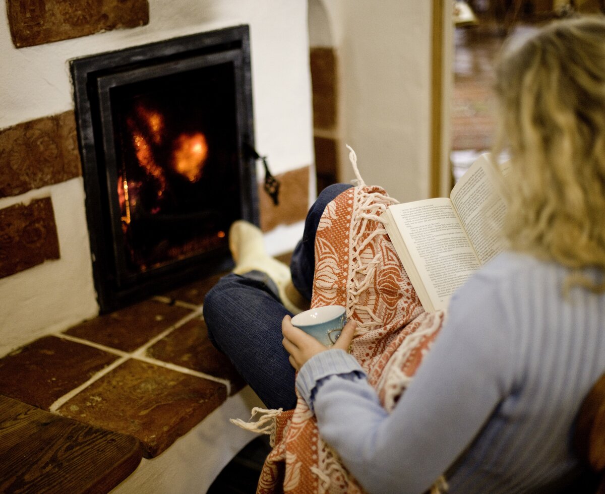 Frau wärmt sich am Ofen auf der Alm | © Urlaub am Bauernhof/ Tom Lamm Eine Person sitzt gemütlich auf dem Boden vor einem Kamin, ein Buch in der Hand. Das warme Feuer bringt ein behagliches Ambiente hervor. | © Urlaub am Bauernhof/ Tom Lamm