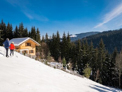 Paar geht zur Hütte | © Michael Stabentheiner Holzhaus in schneebedeckter Berglandschaft, umgeben von verschneiten Tannen und zwei Personen, die am Hang entlanggehen. | © Michael Stabentheiner