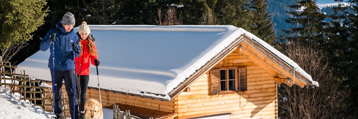 Ein hölzernes Blockhaus umgeben von verschneiter Berglandschaft. Ein Paar in Winterkleidung und ein Hund bewegen sich auf einem Pfad durch den Schnee. Die Sonne scheint auf das Haus und den Winterwald. | © Michael Stabentheiner