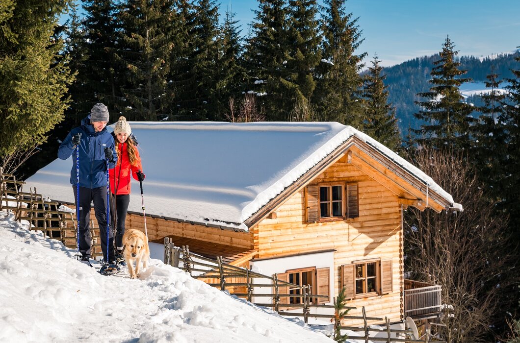 Ein hölzernes Blockhaus umgeben von verschneiter Berglandschaft. Ein Paar in Winterkleidung und ein Hund bewegen sich auf einem Pfad durch den Schnee. Die Sonne scheint auf das Haus und den Winterwald. | © Michael Stabentheiner