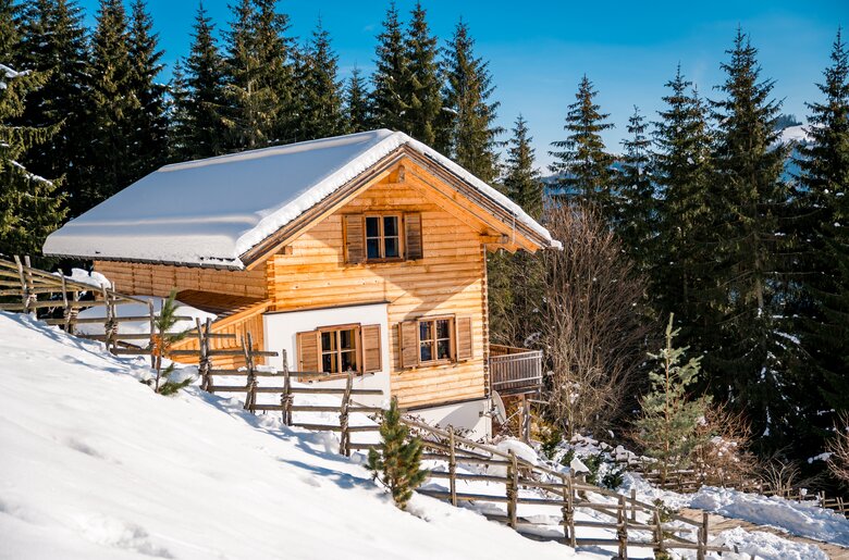 Bergchalet im Winter | © Michael Stabentheiner  Ein kleines, schneebedecktes Holzhaus mit einem Balkon, umgeben von einem Zaun und hohen Tannen in einer winterlichen Landschaft. | © Michael Stabentheiner