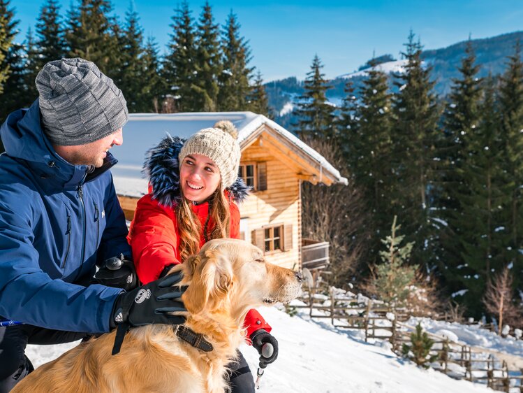 Ein junges Paar in Winterkleidung, das eine Hündin bei einer Holzhütte inmitten verschneiter Wälder und Berge streichelt. Sie lächeln und scheinen die Ruhe und Schönheit der Umgebung zu genießen. | © Michael Stabentheiner