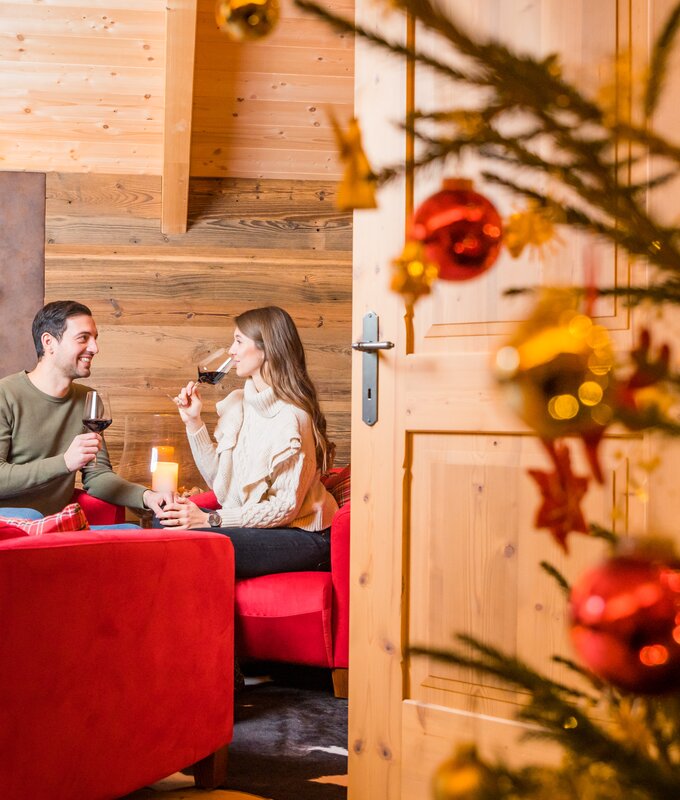 Ein Paar genießt einen gemütlichen Abend in einer festlich geschmückten Holzhütte, während sie Rotwein trinken und sich unterhalten. Weihnachtliche Dekorationen, wie ein Tannenbaum und Christbaumkugeln, verleihen dem Raum eine warme und feierliche Atmosphäre. | © Michael Stabentheiner