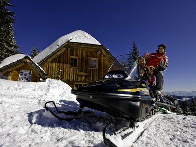 Schneebedecktes, hölzernes Berghaus umgeben von verschneiter Winterlandschaft. Im Vordergrund ein schwarzes Schneemobil, dahinter zwei Personen in Winterbekleidung.