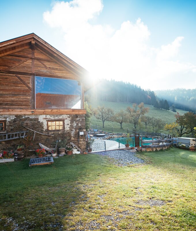 Holzblockhaus inmitten einer idyllischen Berglandschaft mit Wiese, Bäumen und Sträuchern. Das Gebäude verfügt über eine große Glasfront, die den Ausblick in die Natur ermöglicht. Verschiedene Outdoorelemente wie Möbel und Dekorationen lassen auf eine touristische Nutzung des Geländes schließen | © Urlaub am Bauernhof / Daniel Gollner