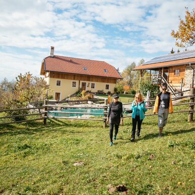 Idyllische Alm mit Holzhaus, umgeben von Herbstlaub und Wiesen. Drei Wanderer schlendern über den Rasen, im Hintergrund ein Pool. Die ländliche Umgebung strahlt Ruhe und Entspannung aus. | © Urlaub am Bauernhof / Daniel Gollner