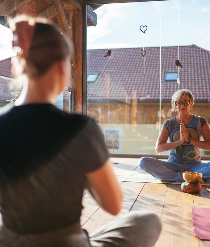 Zwei Frauen in einer ruhigen Umgebung - eine meditiert in Yogastellung, während die andere sie beobachtet. Dekorative Elemente wie Herzen und Vögel hängen an den Wänden, die eine angenehme, entspannende Atmosphäre vermitteln. | © Urlaub am Bauernhof / Daniel Gollner