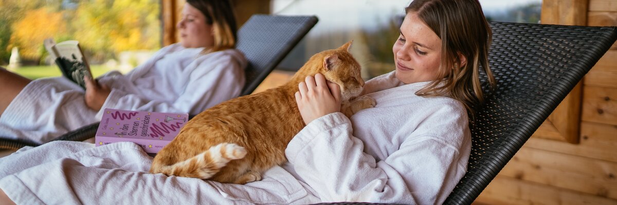 Zwei Frauen entspannen auf Liegestühlen auf der Veranda eines Holzhauses, während eine Katze eine der Frauen begleitet. Die Umgebung ist von Herbstfarben geprägt. | © Urlaub am Bauernhof / Daniel Gollner