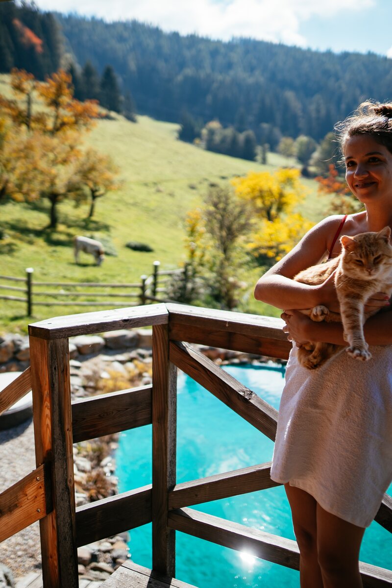 Frau hält eine Katze auf einem hölzernen Balkon über einem Swimmingpool, umgeben von herbstlichen Bäumen und einer bergigen Landschaft. | © Urlaub am Bauernhof / Daniel Gollner