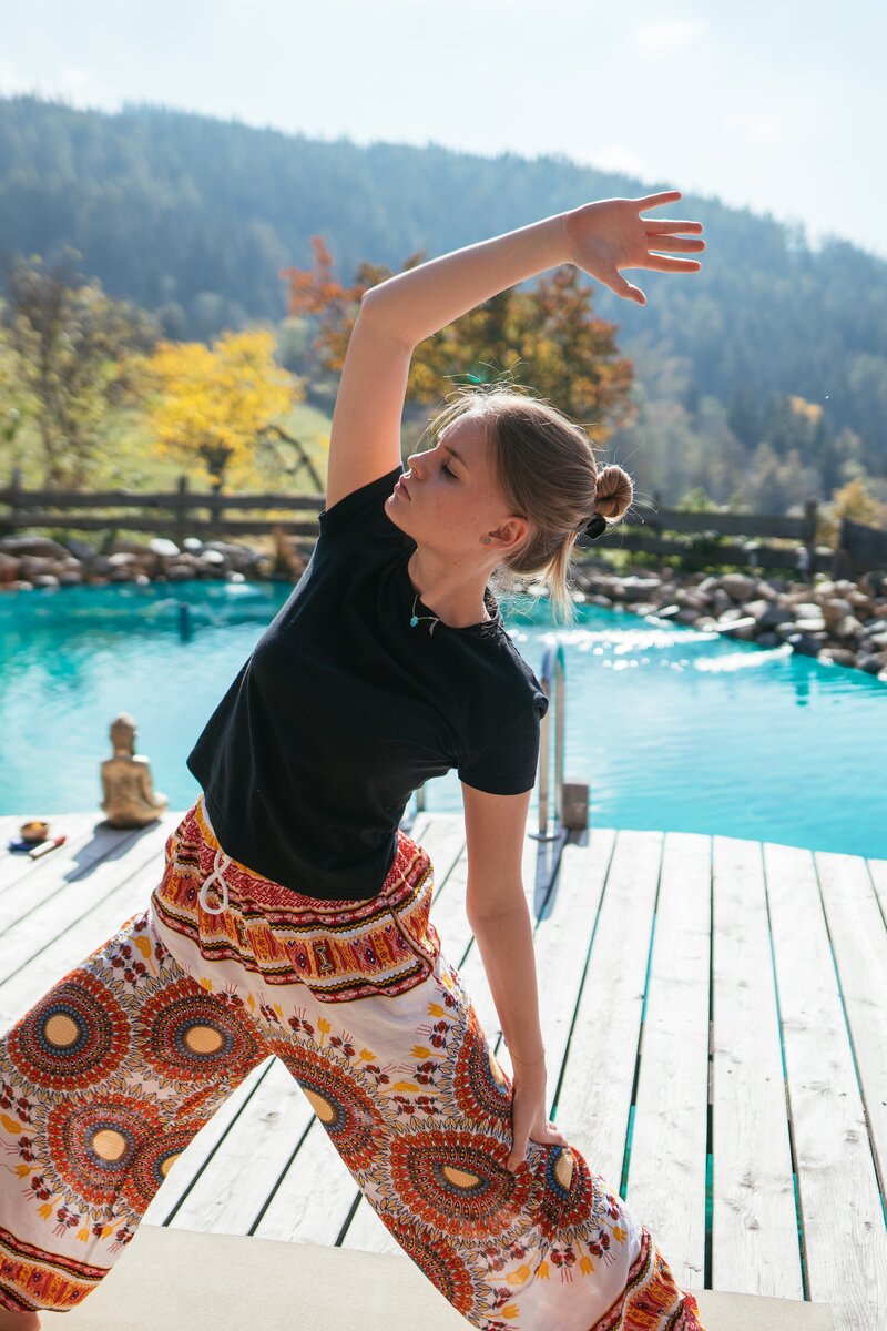 Eine junge Frau übt Yoga-Übungen auf einer Terrasse mit Blick auf einen malerischen See und Berge in herbstlicher Umgebung. Sie trägt bunte, gemusterte Hosen und ein schwarzes Oberteil. Im Hintergrund sind andere Personen zu sehen, die sich ebenfalls entspannen. | © Urlaub am Bauernhof Kärnten / Daniel Gollner