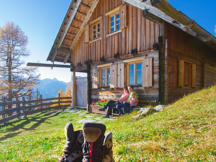 Eine traditionelle Holzhütte in bergiger Umgebung. Zwei Personen sitzen auf einer Bank vor der Hütte, während Wanderschuhe im Vordergrund auf der Wiese liegen.