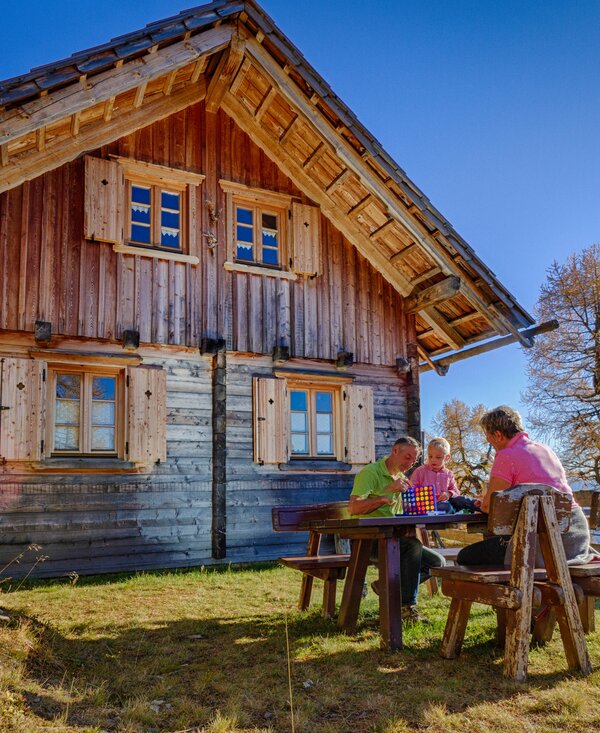 Ein hölzernes Landhaus mit Satteldach und Veranda, umgeben von Bäumen und Wiesen. Eine Gruppe von Personen sitzt an einem rustikalen Tisch und genießt eine ländliche Umgebung.