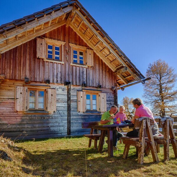 Ein hölzernes Landhaus mit Satteldach und Veranda, umgeben von Bäumen und Wiesen. Eine Gruppe von Personen sitzt an einem rustikalen Tisch und genießt eine ländliche Umgebung.