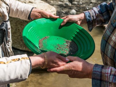 Zwei Männerhände halten gemeinsam eine grüne, rinnenförmige Schürze, in der sich Steine und Sand befinden. Die Oberbekleidung und Armaufschläge deuten auf eine ländliche, bergige Umgebung hin. | © SalzburgerLand Tourismus / Achim Meurer