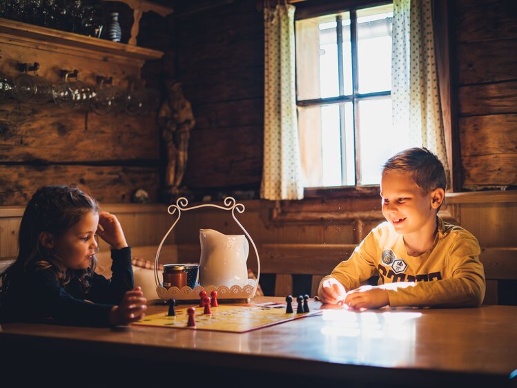 Zwei Kinder spielen Brettspiel in einer urigen Holzhütte mit Vorhängen, Wandregalen und Dekorationen. Der Raum erscheint gemütlich und lädt zum Verweilen ein. | © Urlaub am Bauernhof Salzburg / Daniel Gollner 